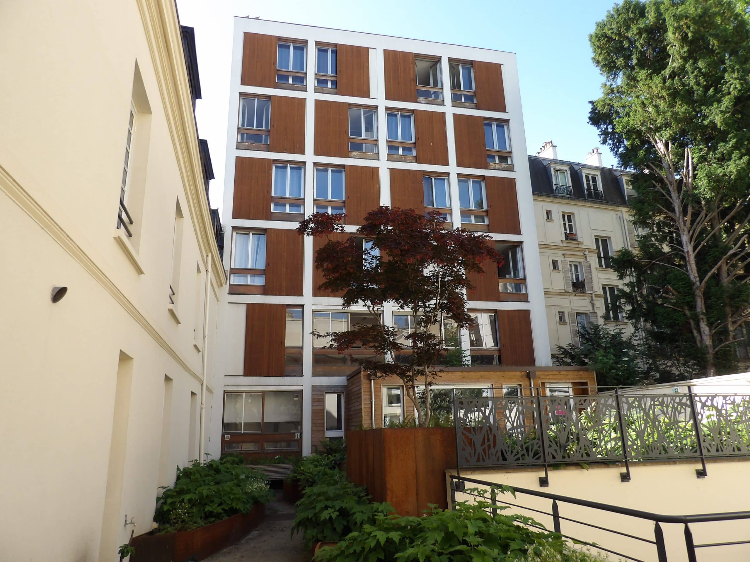 Un immeuble moderne à façade blanche et boiseries se dresse entre un bâtiment crème ancien et un jardin luxuriant avec un arbre rouge et une clôture décorative.