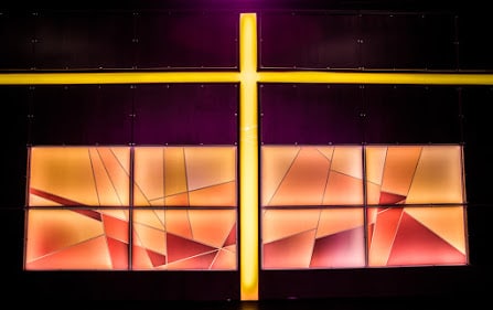 Une grande croix lumineuse jaune coupe un mur sombre. Dessous, huit panneaux affichent des motifs géométriques abstraits, éclairés de teintes chaudes (orange, rouge) sur un fond violet-noir.
