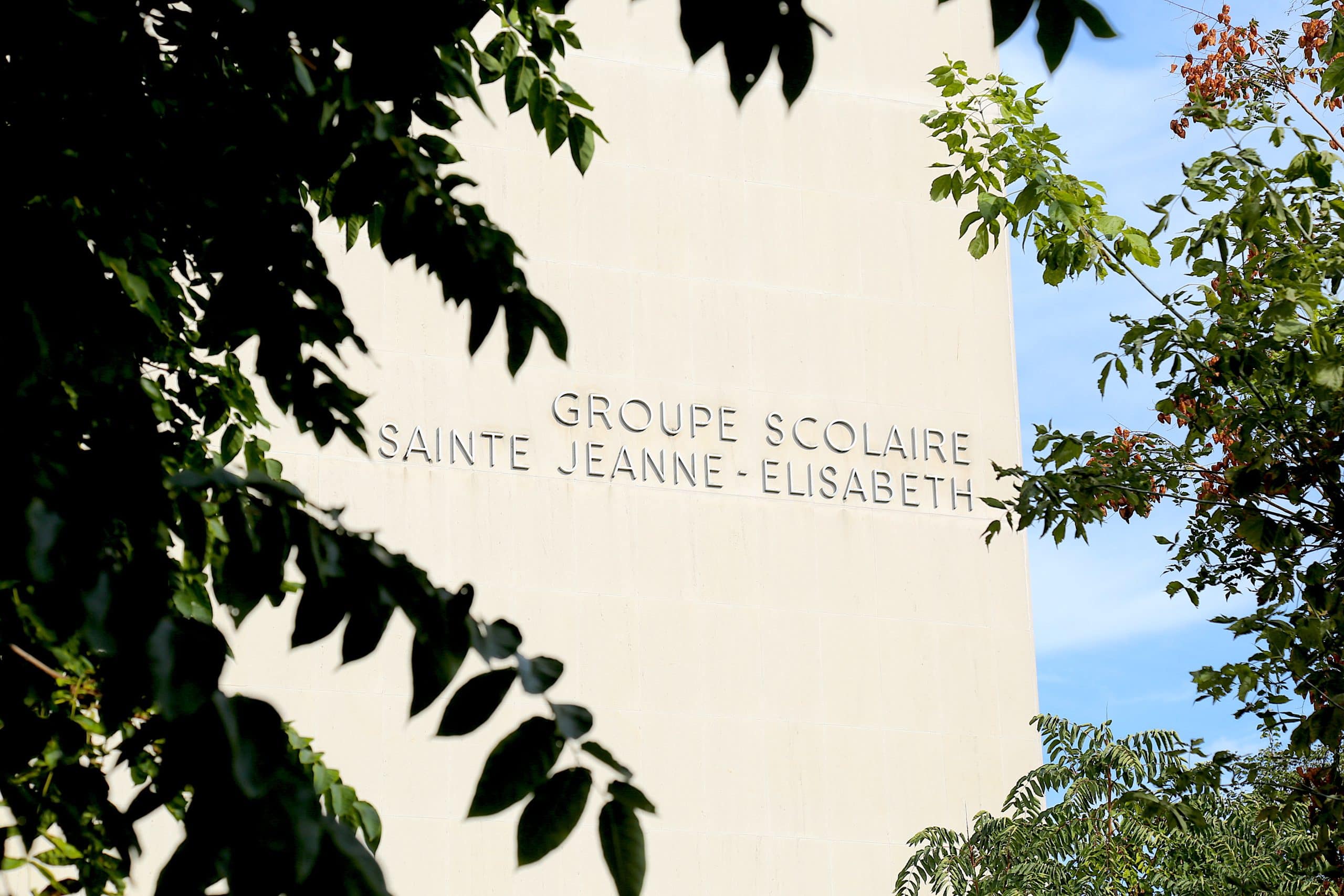 Mur clair d'un bâtiment portant l'inscription "GROUPE SCOLAIRE SAINTE JEANNE - ELISABETH", visible entre des feuillages sombres au premier plan et un ciel bleu.
