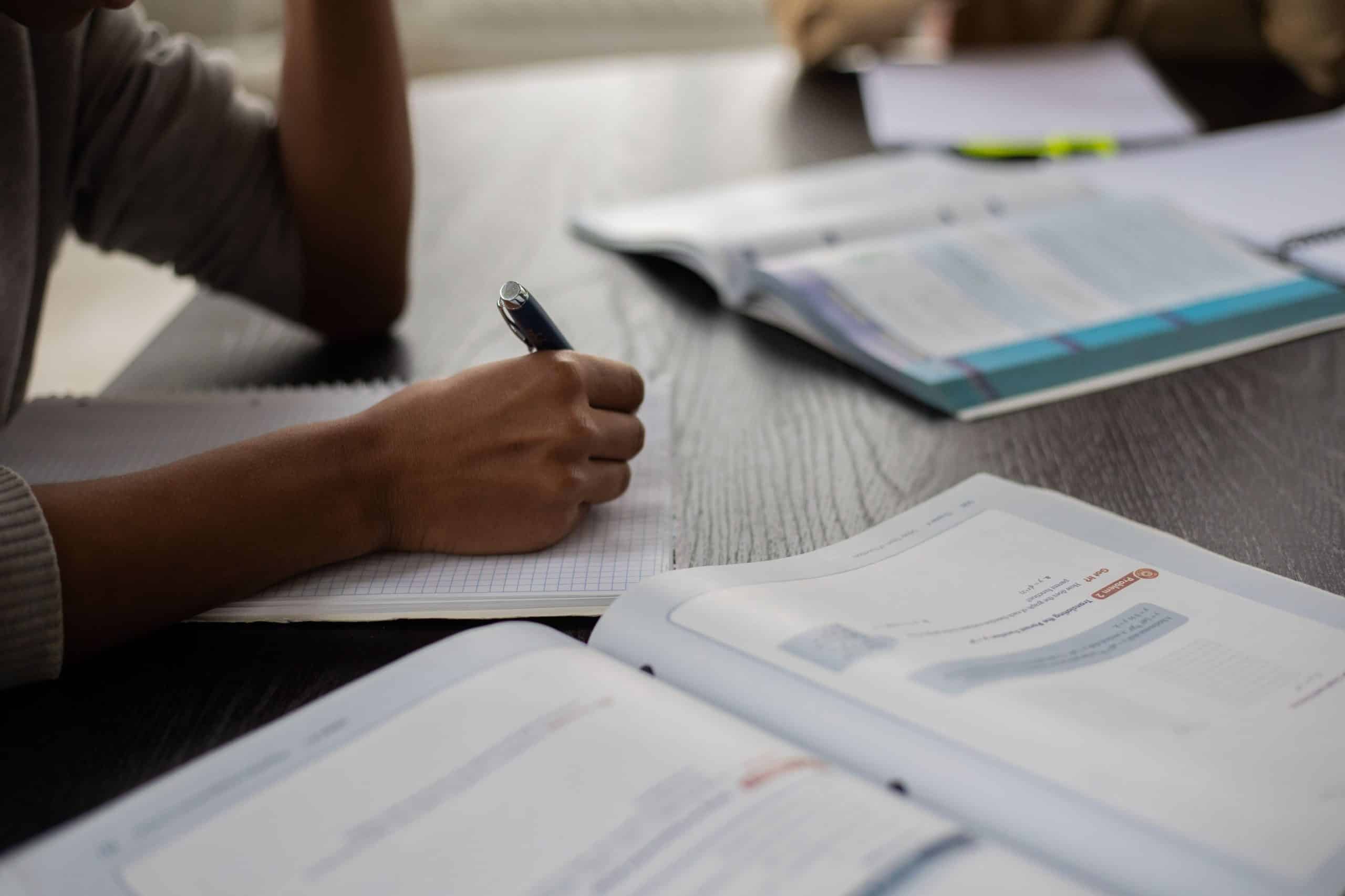 Une main écrit avec un stylo sur un cahier quadrillé, posé sur une table en bois. Des manuels ouverts.