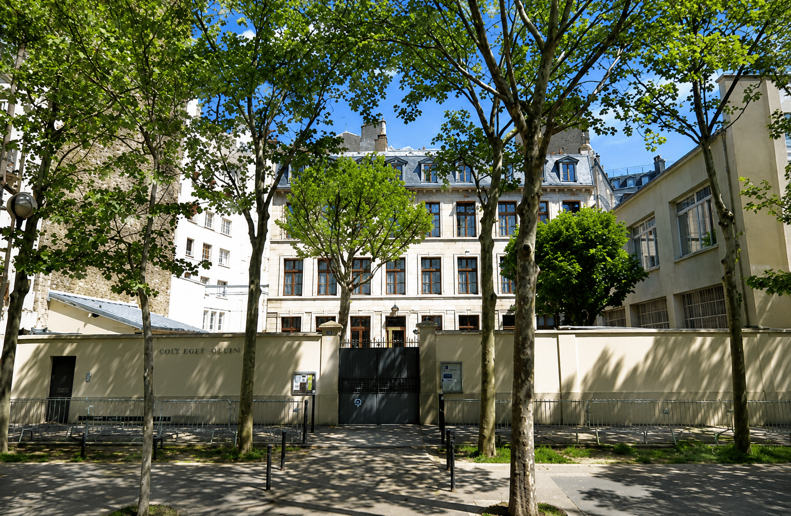 Le bâtiment d'un collège se dresse derrière un mur clair percé d'un grand portail noir, encadré par des arbres feuillus. Le ciel est bleu, le sol ombragé.