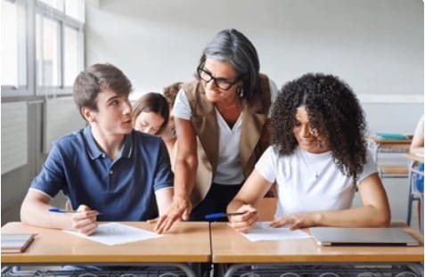 Une professeure aux cheveux gris interagit avec deux élèves assis à leurs pupitres, pointant un document pendant qu'une élève écrit en classe.