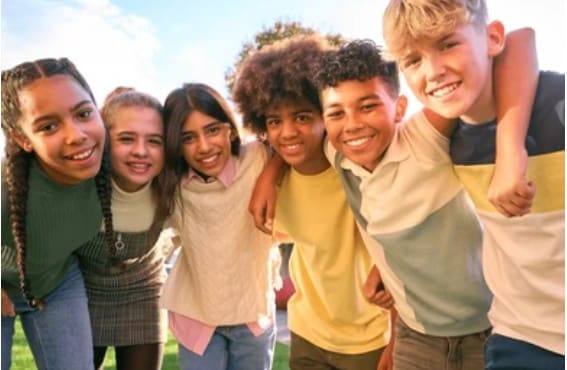 Un groupe d'enfants souriants de diverses ethnies se tient étroitement, bras autour des épaules, en extérieur sous un ciel lumineux avec un arrière-plan verdoyant.