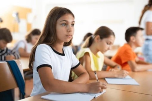 Une jeune fille aux longs cheveux bruns écrit dans un cahier, attentive, assise à un pupitre en classe avec d'autres élèves.