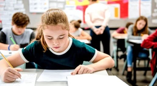 Une jeune fille, concentrée, écrit avec un crayon sur des feuilles. Elle est assise à un pupitre dans une salle de classe avec d'autres élèves et un adulte.